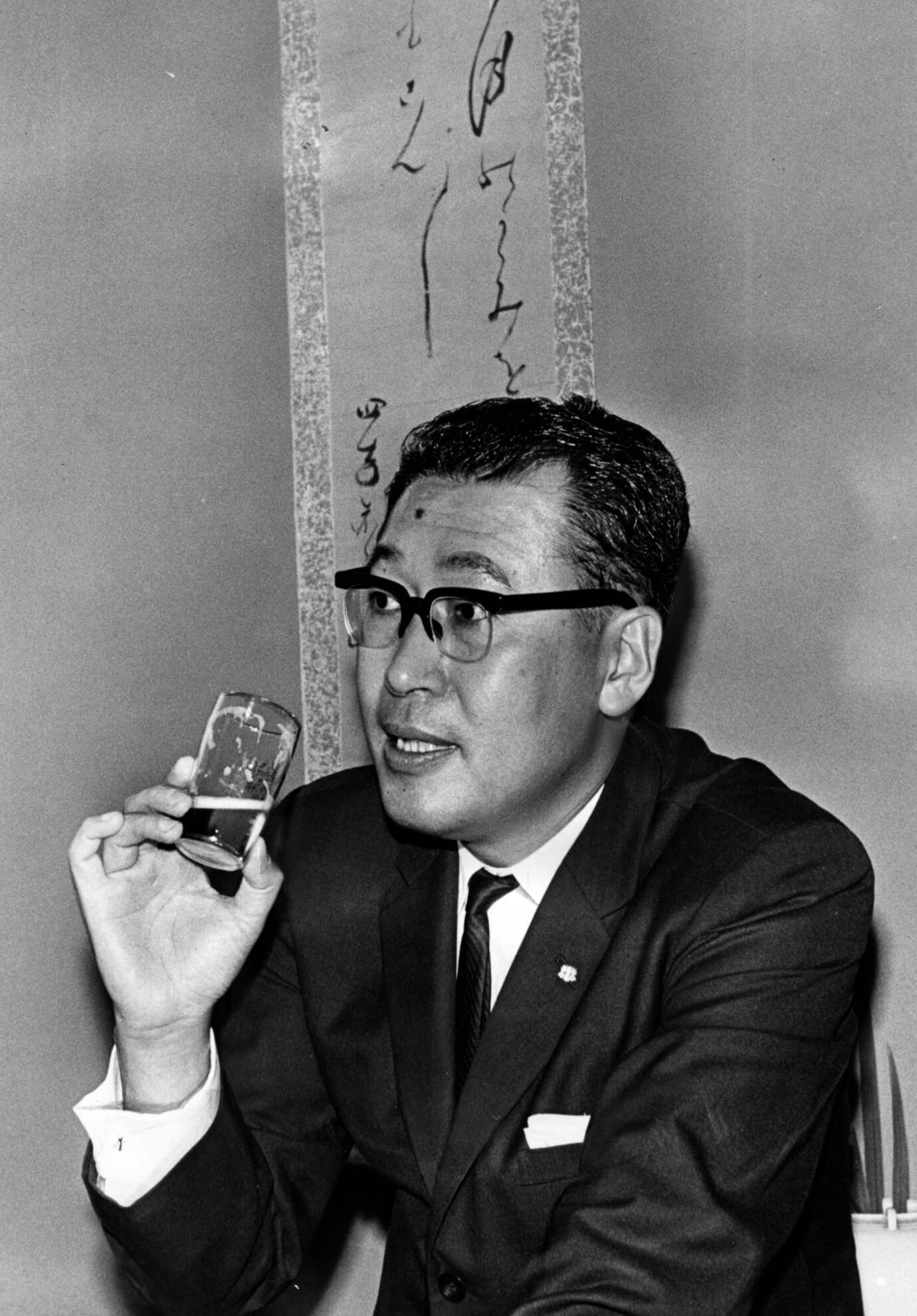 Black-and-white image of Keizo Saji, former Suntory president, with a sample glass of Suntory product