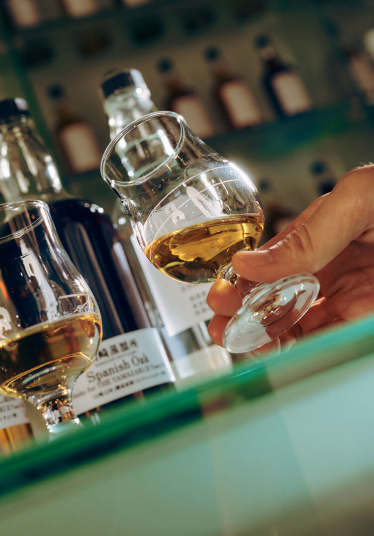 Photo of a hand holding a tasting portion of Suntory product in front of a row of whisky bottles